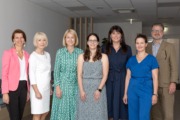 Gruppenfoto von sieben Personen, vier Frauen links, zwei Frauen und ein Mann rechts, die gemeinsam in einem hellen Büroflur stehen und in die Kamera lächeln. Alle sind business-casual gekleidet.