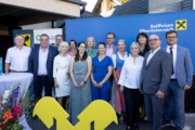 Gruppenfoto mehrerer Frauen und Männer bei einer Veranstaltung von Raiffeisen Oberösterreich. Die Personen stehen im Freien vor blauen und gelben Hintergrundwänden mit dem Raiffeisen-Logo und lächeln in die Kamera. Im Vordergrund ist ein großes gelbes Raiffeisen-Symbol zu sehen.