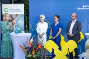 Eine Frau spricht mit einem Mikrofon bei einer Veranstaltung vor Bannern der Österreichischen Gesundheitskasse und Raiffeisen Oberösterreich. Drei weitere Personen stehen daneben und hören zu. Im Vordergrund sind ein Stehtisch mit Blumen und das gelbe Raiffeisen-Logo zu sehen.