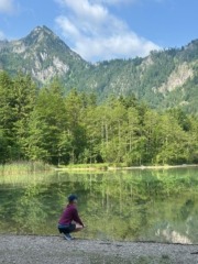 Eine Person in sportlicher Kleidung hockt am Ufer eines klaren Bergsees. Der See spiegelt die umliegenden grünen Bäume und die steilen, bewaldeten Berghänge unter einem leicht bewölkten, blauen Himmel. Die ruhige Naturlandschaft wirkt friedlich und unberührt.