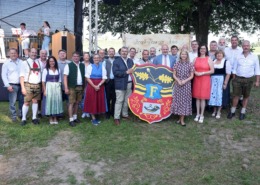 Eine große Gruppe von Menschen steht im Freien vor einer Bühne und einem großen Wappenschild mit dem Buchstaben ‚F‘ und Eichenzweigen. Viele Personen tragen traditionelle Trachten wie Dirndl und Lederhosen, andere sind in moderner Kleidung gekleidet. Im Hintergrund ist eine Musikkapelle mit Blasinstrumenten auf der Bühne zu sehen. Hinter der Gruppe hängt ein Schild mit der Aufschrift ‚Frankenburger Lied‘ und Notenzeilen. Die Szene wirkt wie eine feierliche Veranstaltung oder ein Volksfest in ländlicher Umgebung.