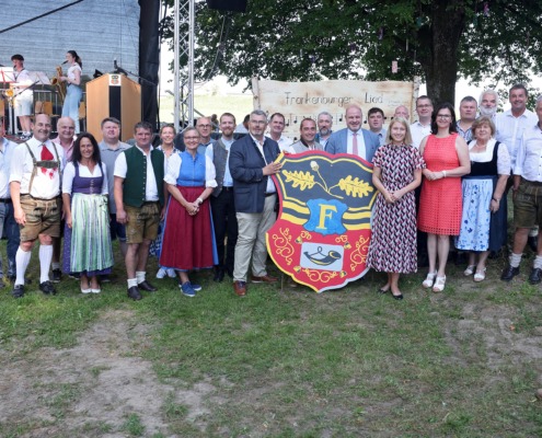 Eine große Gruppe von Menschen steht im Freien vor einer Bühne und einem großen Wappenschild mit dem Buchstaben ‚F‘ und Eichenzweigen. Viele Personen tragen traditionelle Trachten wie Dirndl und Lederhosen, andere sind in moderner Kleidung gekleidet. Im Hintergrund ist eine Musikkapelle mit Blasinstrumenten auf der Bühne zu sehen. Hinter der Gruppe hängt ein Schild mit der Aufschrift ‚Frankenburger Lied‘ und Notenzeilen. Die Szene wirkt wie eine feierliche Veranstaltung oder ein Volksfest in ländlicher Umgebung.