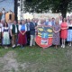 Eine große Gruppe von Menschen steht im Freien vor einer Bühne und einem großen Wappenschild mit dem Buchstaben ‚F‘ und Eichenzweigen. Viele Personen tragen traditionelle Trachten wie Dirndl und Lederhosen, andere sind in moderner Kleidung gekleidet. Im Hintergrund ist eine Musikkapelle mit Blasinstrumenten auf der Bühne zu sehen. Hinter der Gruppe hängt ein Schild mit der Aufschrift ‚Frankenburger Lied‘ und Notenzeilen. Die Szene wirkt wie eine feierliche Veranstaltung oder ein Volksfest in ländlicher Umgebung.