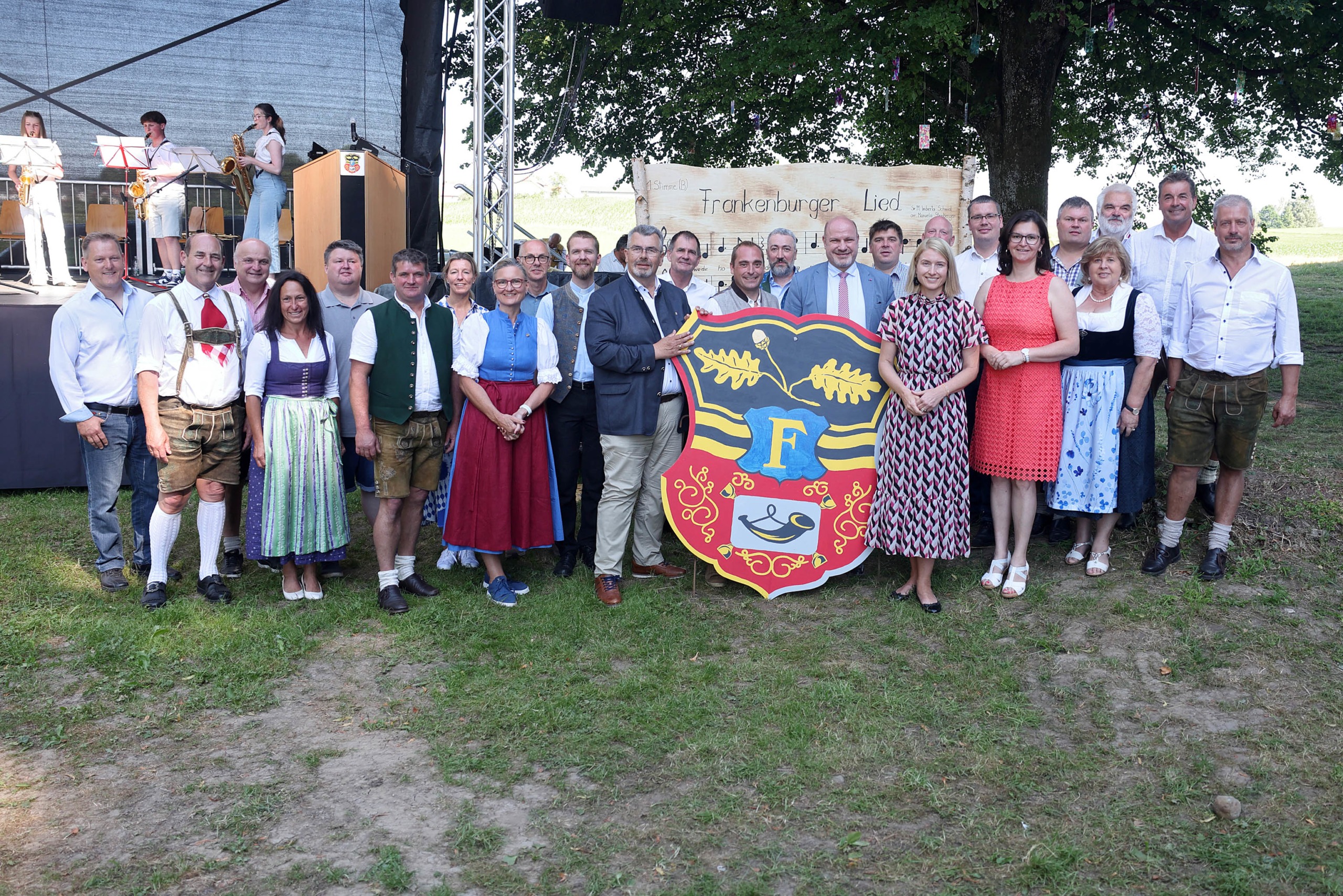 Eine große Gruppe von Menschen steht im Freien vor einer Bühne und einem großen Wappenschild mit dem Buchstaben ‚F‘ und Eichenzweigen. Viele Personen tragen traditionelle Trachten wie Dirndl und Lederhosen, andere sind in moderner Kleidung gekleidet. Im Hintergrund ist eine Musikkapelle mit Blasinstrumenten auf der Bühne zu sehen. Hinter der Gruppe hängt ein Schild mit der Aufschrift ‚Frankenburger Lied‘ und Notenzeilen. Die Szene wirkt wie eine feierliche Veranstaltung oder ein Volksfest in ländlicher Umgebung.