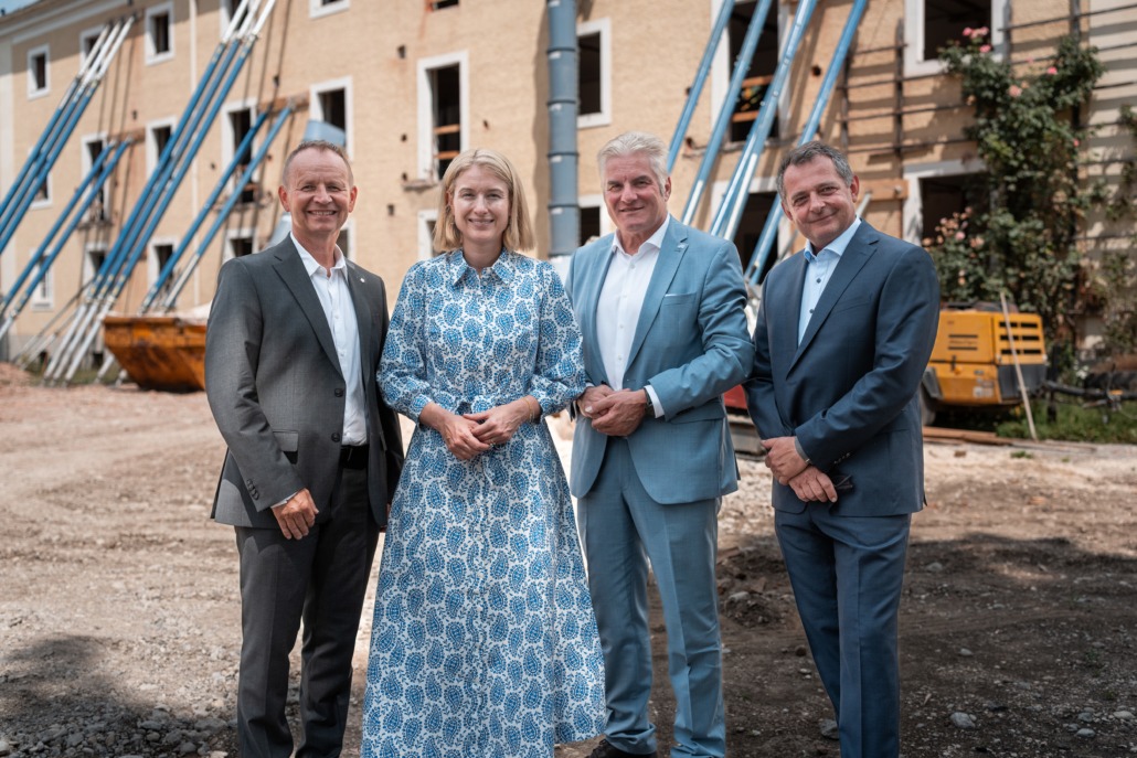 Vier Personen in eleganter Kleidung stehen nebeneinander auf einer Baustelle vor einem alten Gebäude mit gelber Fassade und offenen Fensteröffnungen. Das Gebäude wird mit blauen Stahlträgern abgestützt. Im Hintergrund sind Bauschutt, ein gelber Container und Baumaschinen zu sehen. Die Personen lächeln in die Kamera – die Szene wirkt wie eine offizielle Besichtigung oder ein Pressetermin im Rahmen eines Bauprojekts.