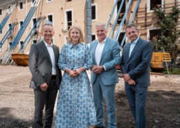 Vier Personen in eleganter Kleidung stehen nebeneinander auf einer Baustelle vor einem alten Gebäude mit gelber Fassade und offenen Fensteröffnungen. Das Gebäude wird mit blauen Stahlträgern abgestützt. Im Hintergrund sind Bauschutt, ein gelber Container und Baumaschinen zu sehen. Die Personen lächeln in die Kamera – die Szene wirkt wie eine offizielle Besichtigung oder ein Pressetermin im Rahmen eines Bauprojekts.