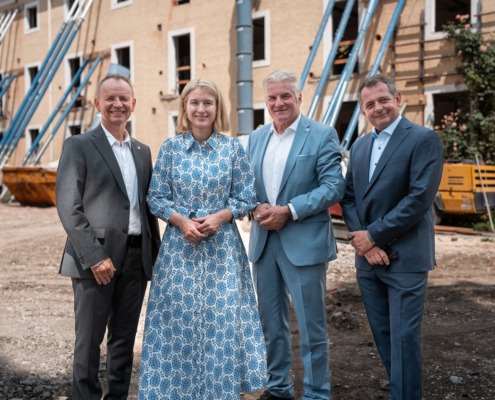 Vier Personen in eleganter Kleidung stehen nebeneinander auf einer Baustelle vor einem alten Gebäude mit gelber Fassade und offenen Fensteröffnungen. Das Gebäude wird mit blauen Stahlträgern abgestützt. Im Hintergrund sind Bauschutt, ein gelber Container und Baumaschinen zu sehen. Die Personen lächeln in die Kamera – die Szene wirkt wie eine offizielle Besichtigung oder ein Pressetermin im Rahmen eines Bauprojekts.