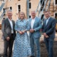 Vier Personen in eleganter Kleidung stehen nebeneinander auf einer Baustelle vor einem alten Gebäude mit gelber Fassade und offenen Fensteröffnungen. Das Gebäude wird mit blauen Stahlträgern abgestützt. Im Hintergrund sind Bauschutt, ein gelber Container und Baumaschinen zu sehen. Die Personen lächeln in die Kamera – die Szene wirkt wie eine offizielle Besichtigung oder ein Pressetermin im Rahmen eines Bauprojekts.