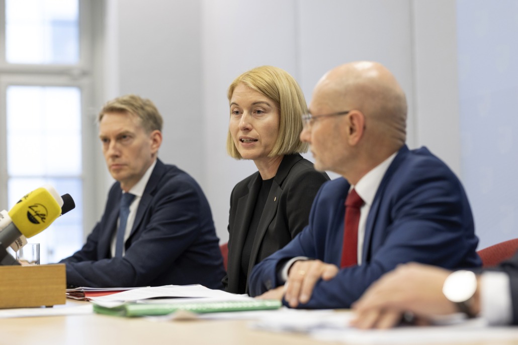 Drei Personen sitzen bei einer Pressekonferenz am Tisch und sprechen.