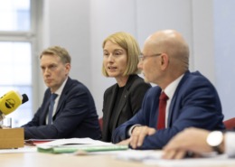 Drei Personen sitzen bei einer Pressekonferenz am Tisch und sprechen.