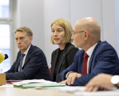 Drei Personen sitzen bei einer Pressekonferenz am Tisch und sprechen.