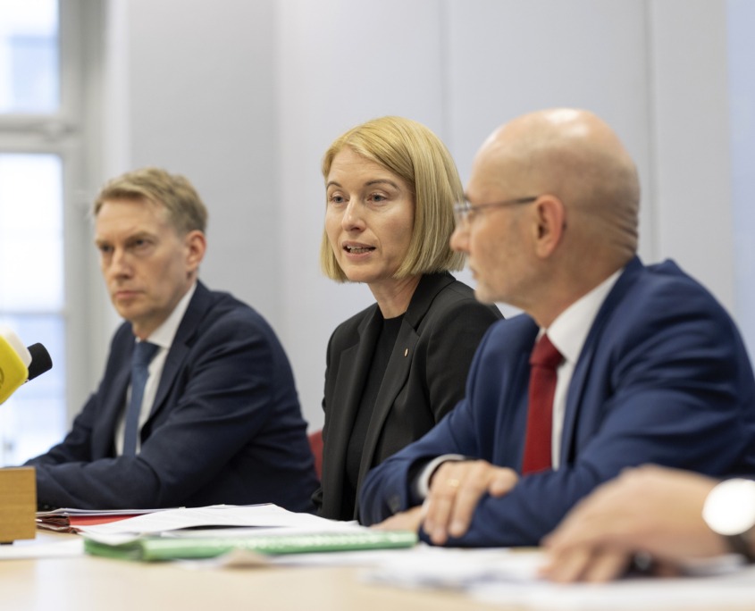 Drei Personen sitzen bei einer Pressekonferenz am Tisch und sprechen.