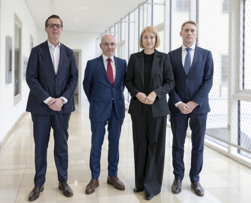 Vier Personen in dunklen Business-Outfits stehen in einem hellen Gang.