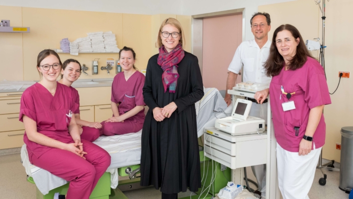 Sechs Personen aus dem Gesundheitsbereich stehen in einem Behandlungsraum neben einem Krankenbett und medizinischen Geräten.