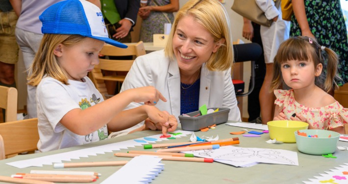 Zwei Kinder basteln mit Papier und Stiften an einem Tisch, eine Erwachsene sitzt daneben.